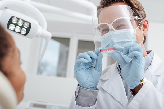 Dentist wearing protective gear holding up a denture to patient